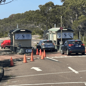A road with 2 cars stopped at small buildings with boom gates. One road is blocked off with orange witch's hats. Thick gumtrees are visible behind the entry stations