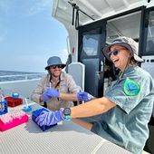 Two people in green shirts and widebrim hats sitting on the back of a boat at a desk covered in medical sample vials